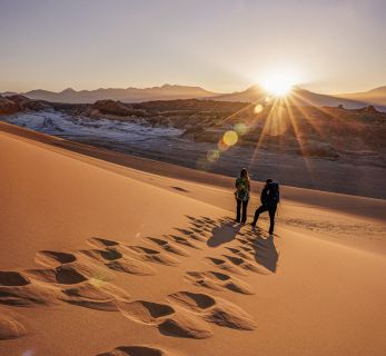 Travelers hiking atop golden sand dunes at sunrise in a remote desert landscape, with panoramic mountain views and sun flares enhancing the scene.