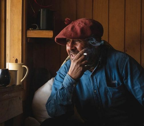 Gaucho in a red beret and denim shirt smiling while smoking, seated in a cozy wooden cabin in Torres del Paine.