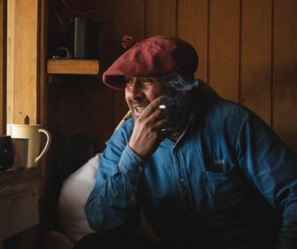Gaucho in a red beret and denim shirt smiling while smoking, seated in a cozy wooden cabin in Torres del Paine.