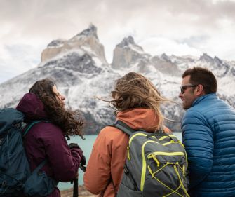 Three hikers with backpacks enjoying a scenic view of snow-capped mountains and a turquoise lake in Patagonia.