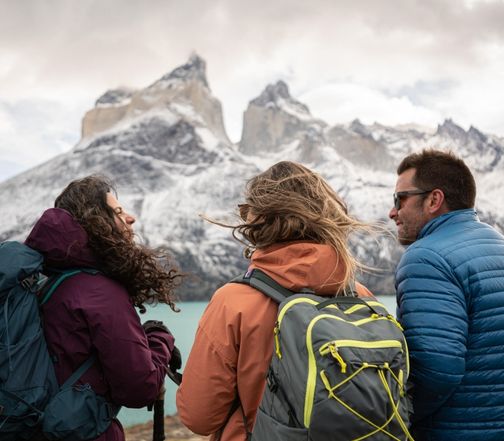Three hikers with backpacks enjoying a scenic view of snow-capped mountains and a turquoise lake in Patagonia.
