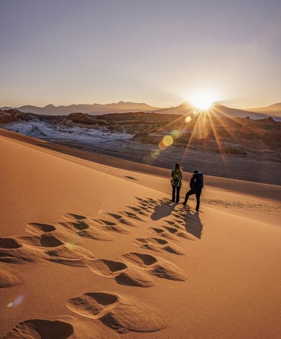 Travelers hiking atop golden sand dunes at sunrise in a remote desert landscape, with panoramic mountain views and sun flares enhancing the scene.