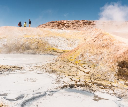 Steaming mineral pools in a rocky desert with visitors on a ridge.
