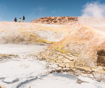 Steaming mineral pools in a rocky desert with visitors on a ridge.