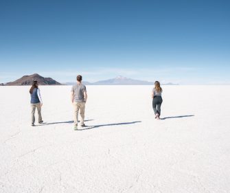 Three people walking across a vast white salt flat toward distant mountains.