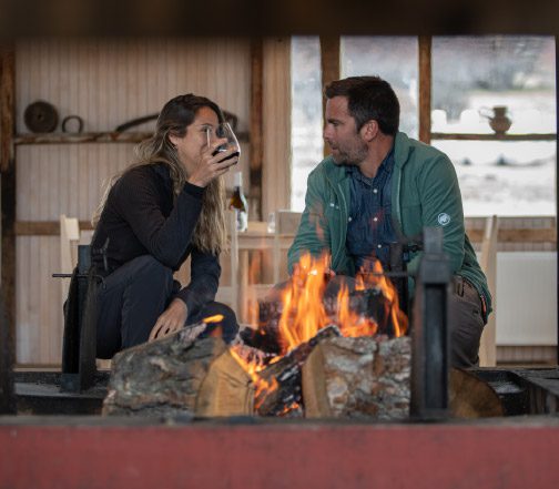 A group of people are seated around a table inside a rustic wooden dining space with large windows, sharing food and drinks while looking out toward a wide, mountainous landscape in the distance.