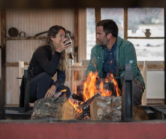 A group of people are seated around a table inside a rustic wooden dining space with large windows, sharing food and drinks while looking out toward a wide, mountainous landscape in the distance.