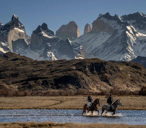 People on horseback cross a shallow river in an open plain, with Macizo Paine in the background