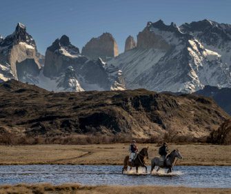 People on horseback cross a shallow river in an open plain, with Macizo Paine in the background