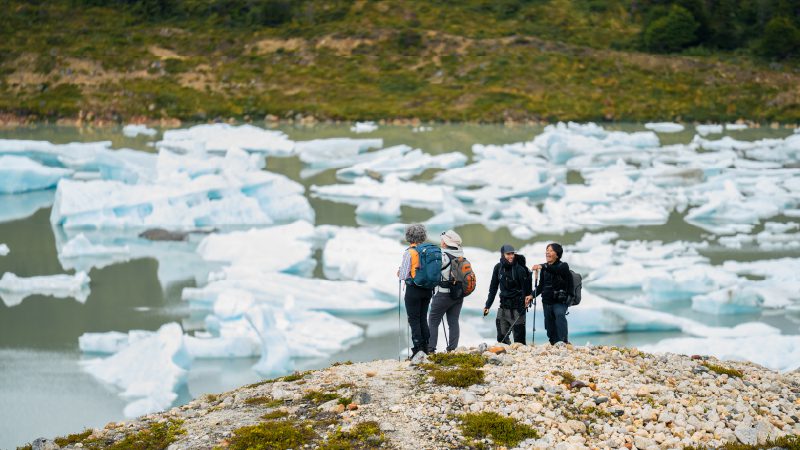 A group of hikers stands on a rocky ridge overlooking a lake filled with floating ice chunks.