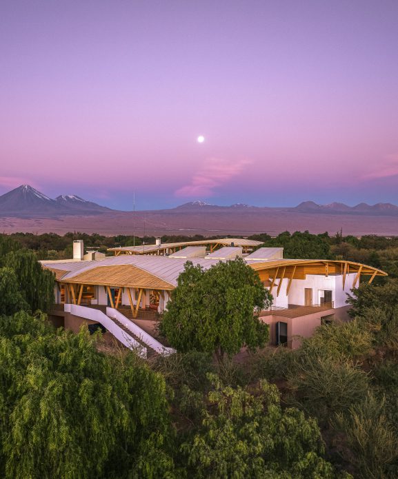 Explora Atacama luxury lodge at dusk, surrounded by desert vegetation, with Andean mountains and a soft purple sky in northern Chile.
