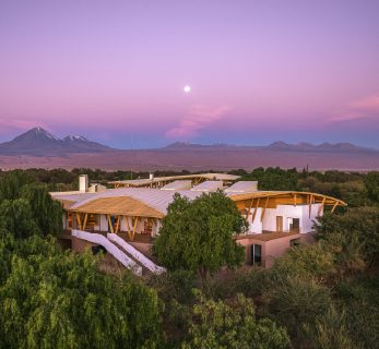 Explora Atacama luxury lodge at dusk, surrounded by desert vegetation, with Andean mountains and a soft purple sky in northern Chile.
