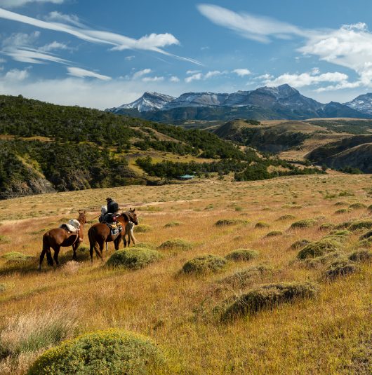 http://Two%20riders%20on%20horseback%20crossing%20an%20open%20grassland%20with%20mountains%20in%20the%20distance.
