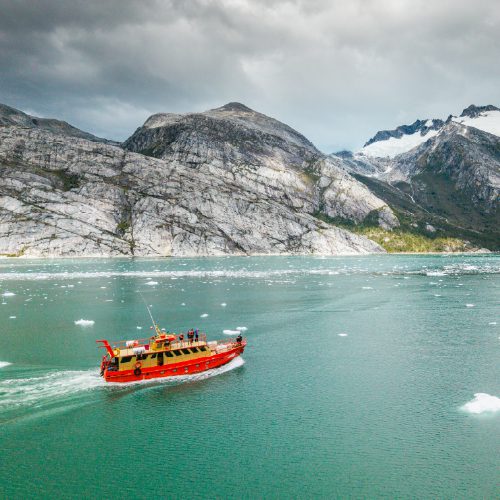 A red boat cruises across icy turquoise water near rocky mountains.