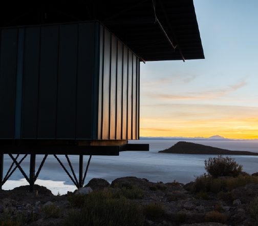 A modern building elevated on metal supports overlooks a vast salt flat or lake, with low vegetation in the foreground and distant islands and mountains glowing under a soft sunset sky.