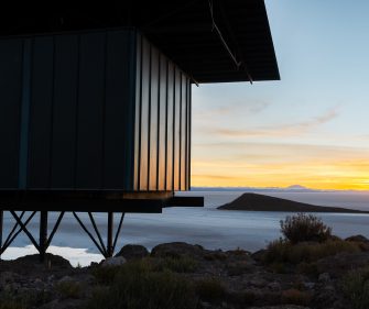 A modern building elevated on metal supports overlooks a vast salt flat or lake, with low vegetation in the foreground and distant islands and mountains glowing under a soft sunset sky.