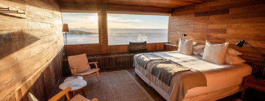 A warm wooden bedroom with a large bed and wide windows overlooking water and distant mountains.
