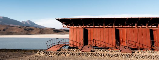 A long red wooden building stands beside a calm lake in a barren desert landscape, with mountains in the background under a clear blue sky.