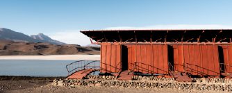 A long red wooden building stands beside a calm lake in a barren desert landscape, with mountains in the background under a clear blue sky.