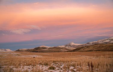 A wide open landscape with dry grass and light patches of snow, leading to distant hills and mountains under a soft pink and orange sunset sky at Torres del Paine Conservation Reserve