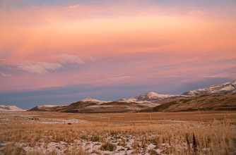 A wide open landscape with dry grass and light patches of snow, leading to distant hills and mountains under a soft pink and orange sunset sky at Torres del Paine Conservation Reserve