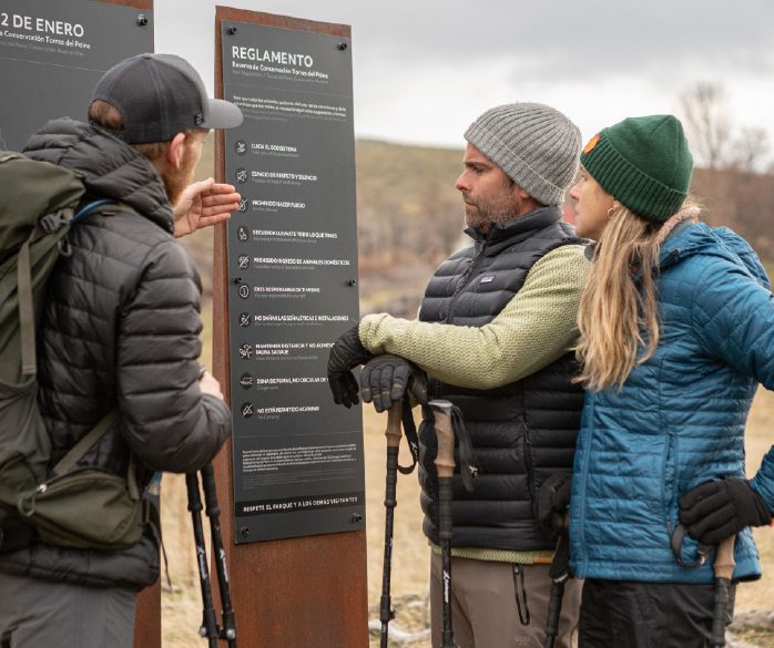 A small group of hikers stands outdoors at a nature reserve, wearing cold‑weather clothing and holding trekking poles while reading and discussing a posted set of park rules on an informational sign.