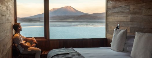 A bedroom interior with a large window facing a lake and mountains.