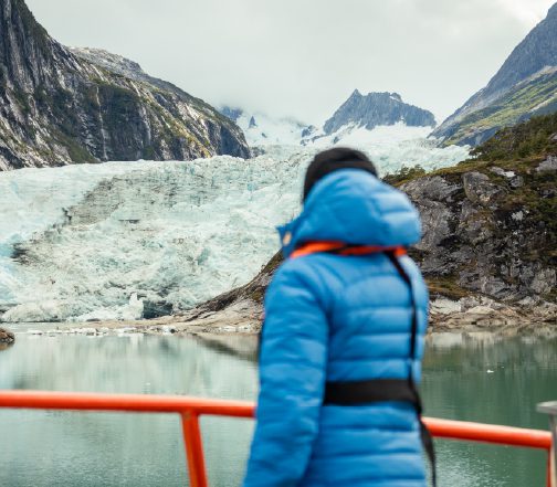 A person wearing a blue jacket stands on a viewing platform, looking out over a calm lake toward a massive glacier surrounded by steep, rocky mountains under a cloudy sky.