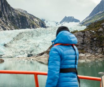 A person wearing a blue jacket stands on a viewing platform, looking out over a calm lake toward a massive glacier surrounded by steep, rocky mountains under a cloudy sky.