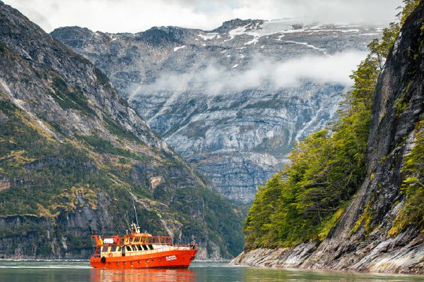 A red boat floats on calm water between steep, rocky mountains with mist and clouds overhead.
