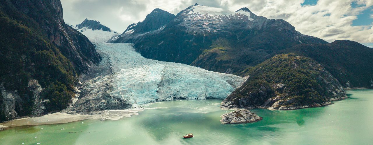 http://A%20large%20glacier%20descends%20between%20dark%20mountains%20into%20a%20milky%20turquoise%20lake,%20where%20a%20small%20boat%20floats%20near%20the%20ice%20under%20a%20cloudy%20sky.