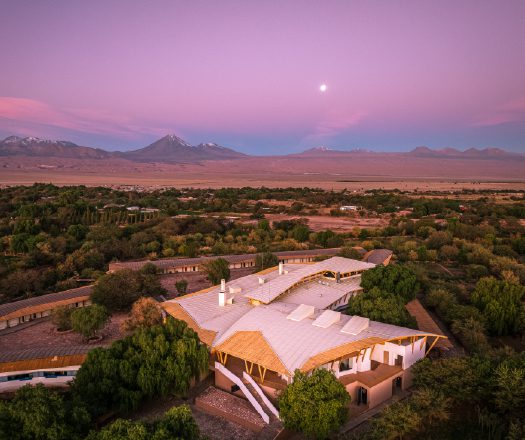 An aerial view of a lodge complex set among green trees in a desert landscape, with distant mountains under a pink and purple twilight sky.