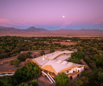 An aerial view of a lodge complex set among green trees in a desert landscape, with distant mountains under a pink and purple twilight sky.