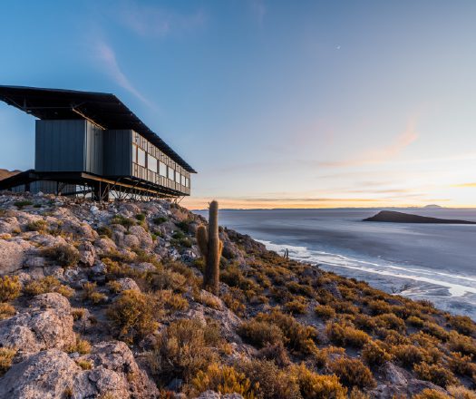 A modern building sits on a rocky hillside overlooking a vast lake or salt flat, surrounded by sparse desert vegetation and lit by warm sunset light.