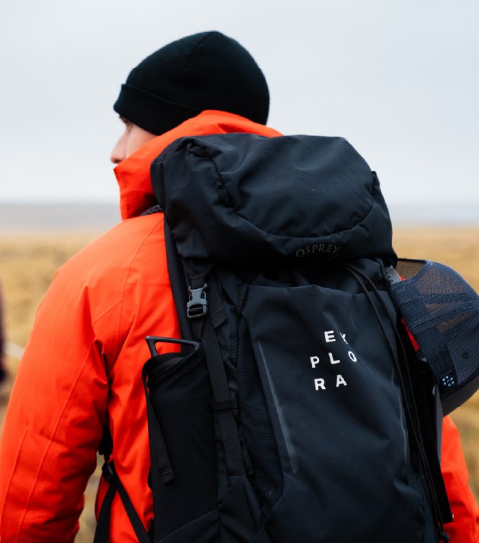 A traveler in an orange jacket carries a large backpack in an open landscape.