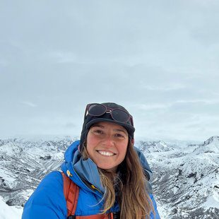 Expeditions Experience Manager at Explora, smiling at high altitude with a vast snow-covered mountain range behind her, wearing a blue Explora down jacket and mountain gear