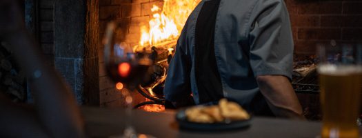Chef cooking over an open fire in a Patagonian restaurant with local food and craft beer in the foreground