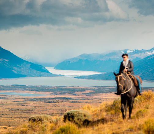 Gaucho on horseback riding across golden Patagonian steppe with Lago Argentino and a distant glacier visible in the valley below, El Calafate, Argentina