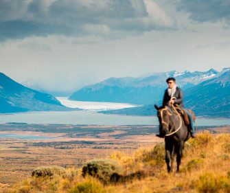 Gaucho on horseback riding across golden Patagonian steppe with Lago Argentino and a distant glacier visible in the valley below, El Calafate, Argentina