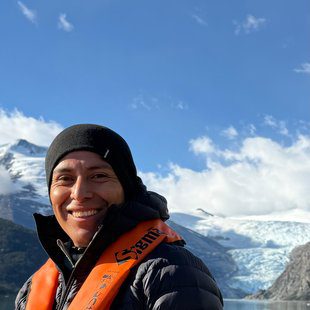 Sacred Mountains Expedition Coordinator at Explora, smiling aboard a boat with a glacier and turquoise lake in the background, wearing a black beanie and orange life vest
