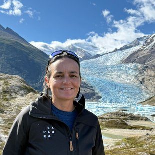Expeditions Coordinator at Explora, smiling in front of a Patagonian glacier with snow-capped mountains, wearing an Explora softshell jacket and sunglasses on her head