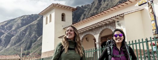 Two travelers walk near a historic church in the Sacred Valley of Peru, framed by dramatic Andean mountains and colonial architecture.