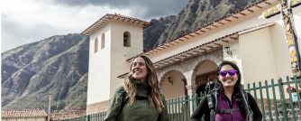 Two travelers walk near a historic church in the Sacred Valley of Peru, framed by dramatic Andean mountains and colonial architecture.