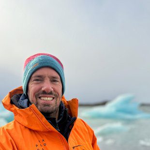 Expeditions Operations Manager at Explora, smiling in a bright orange jacket and colorful beanie in front of a glacier lagoon with floating icebergs