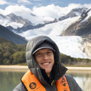 Expeditions Sales Manager at Explora, smiling outdoors in Patagonia with a glacier and snow-capped mountains in the background, wearing a hooded down jacket and life vest