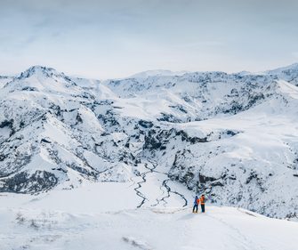 Two explorers overlooking a snow-covered volcanic mountain range and frozen river in Iceland's Winter wilderness
 