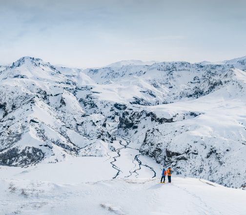 Two explorers overlooking a snow-covered volcanic mountain range and frozen river in Iceland's Winter wilderness